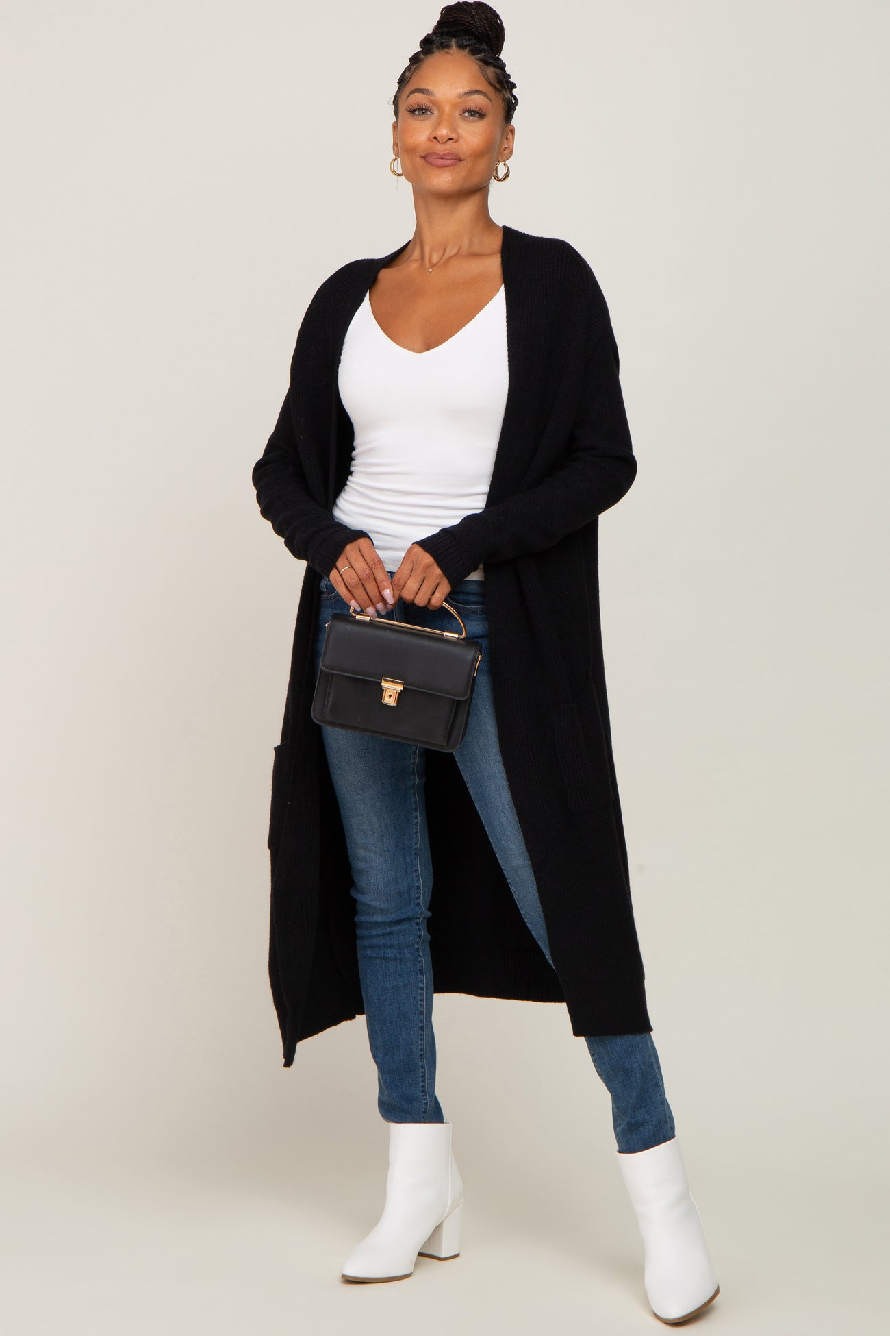 woman wearing long black cardigan white top blue jeans and white boots holding a black handbag