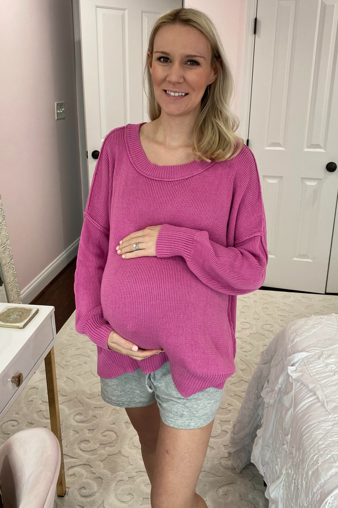 woman wearing a bright pink oversized maternity sweater and grey shorts, holding her baby bump