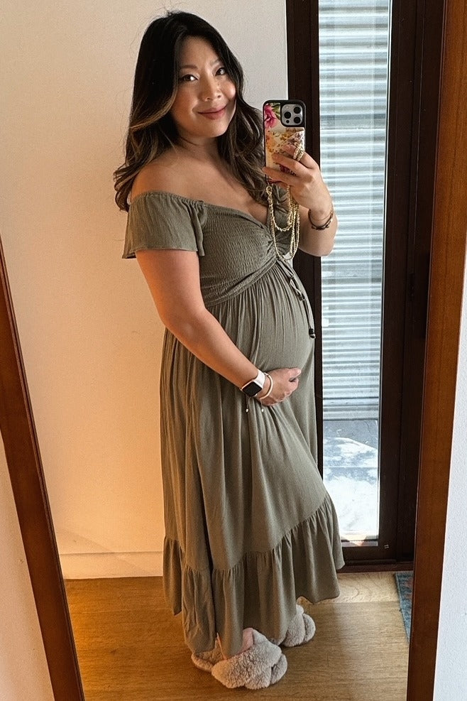 woman wearing a light olive green off-shoulder maternity dress with a tiered skirt and holding her baby bump