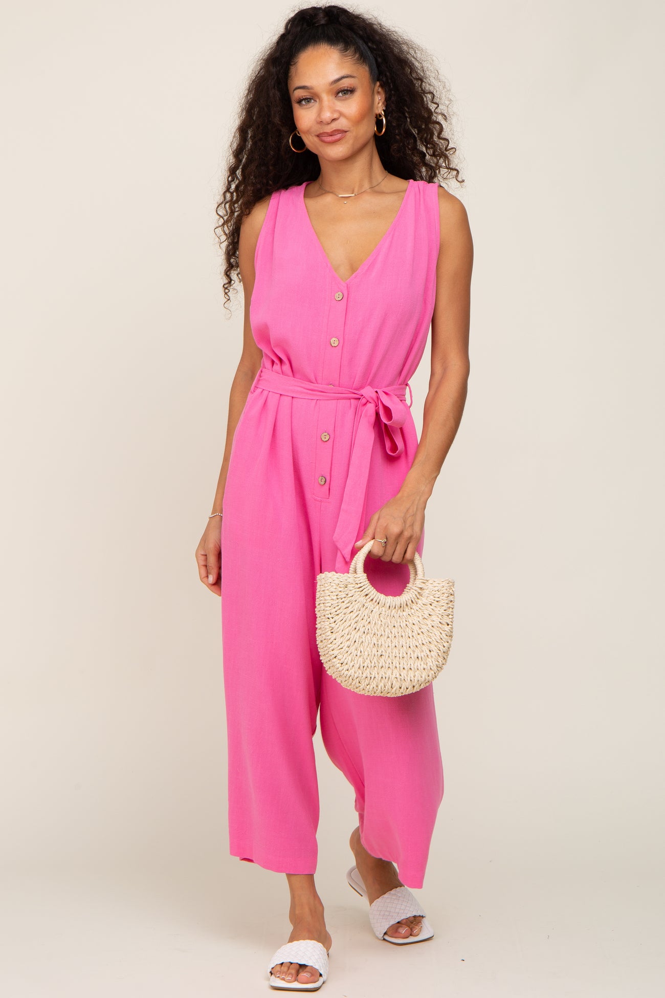 woman wearing a pink sleeveless jumpsuit with a tie waist and buttons, holding a natural woven handbag