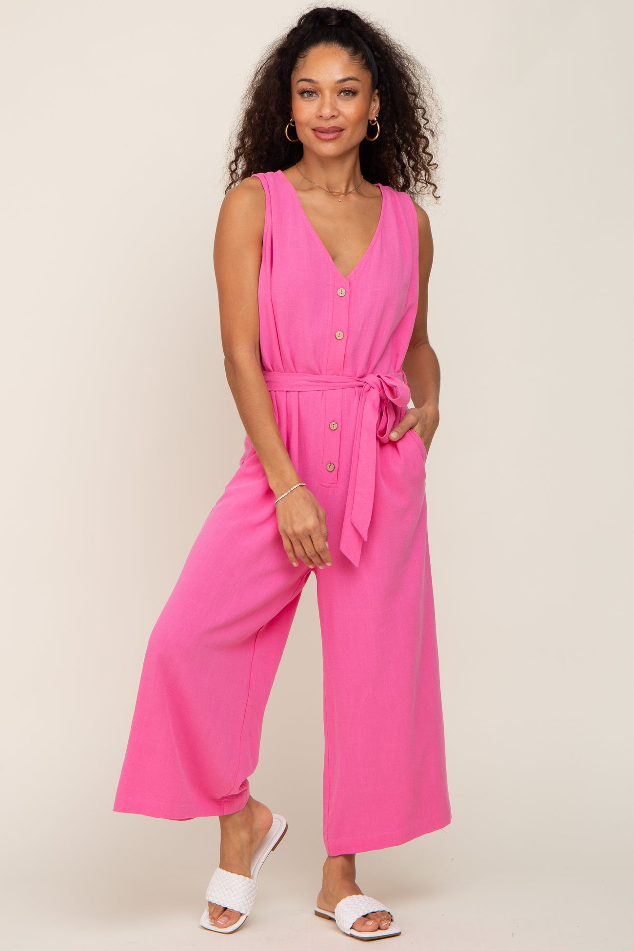 woman wearing a bright pink sleeveless jumpsuit with a tie belt and button details, styled with white slide sandals