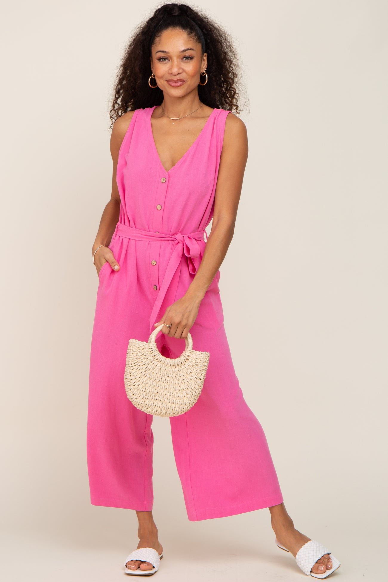 woman wearing a bright pink sleeveless jumpsuit with buttons and a tie waist, holding a woven handbag