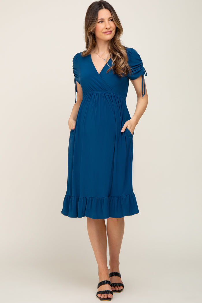 woman wearing a teal maternity dress with puff sleeves, a ruffled hem, and pockets, showing her baby bump
