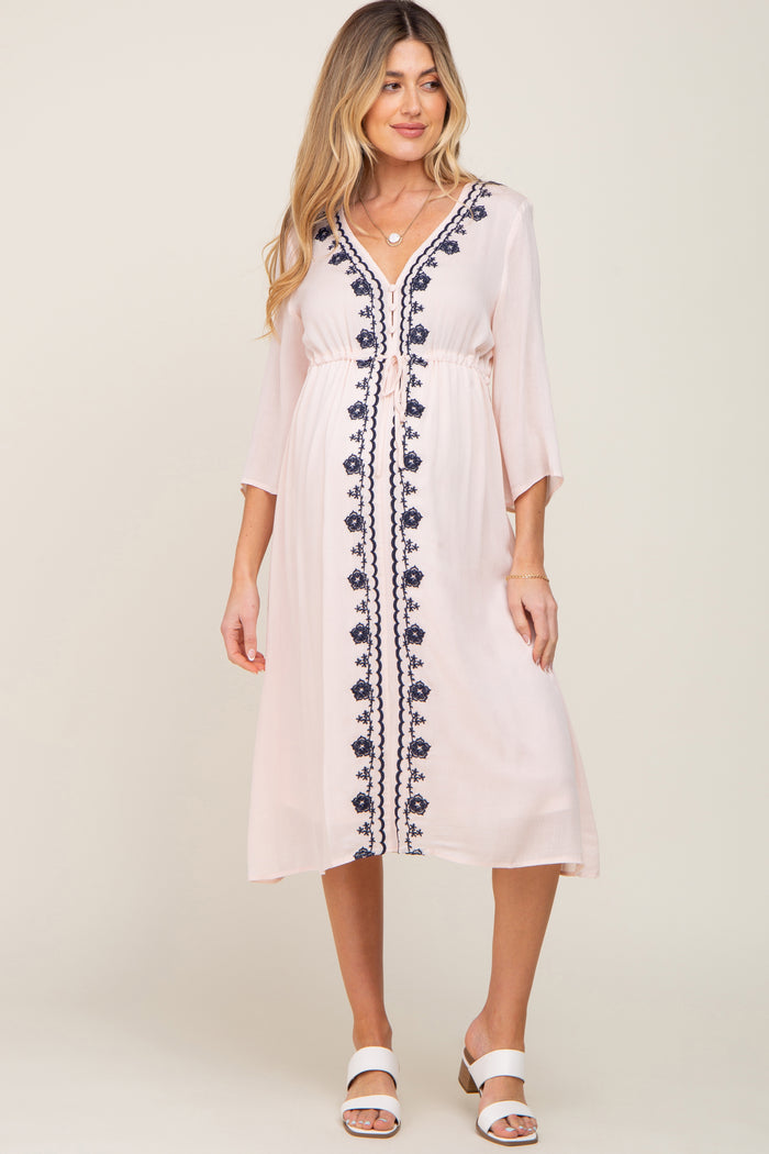 woman wearing a light pink maternity dress with navy embroidery and three-quarter sleeves, standing and smiling