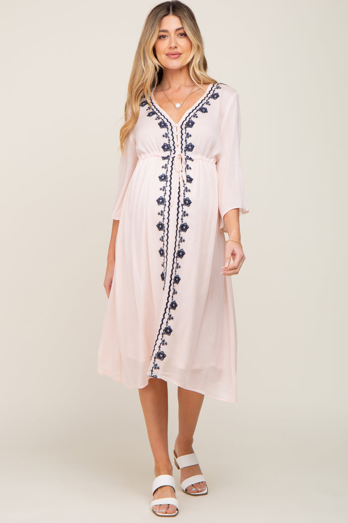 woman wearing a light pink maternity dress with navy floral embroidery and a tie waist detail