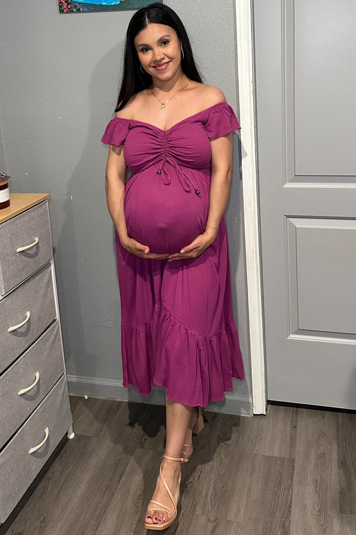 smiling pregnant woman in a purple off-the-shoulder maternity dress holding her baby bump