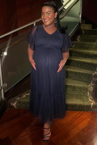 woman wearing a navy blue maternity gown with short tulle sleeves and a flowing skirt, holding her baby bump