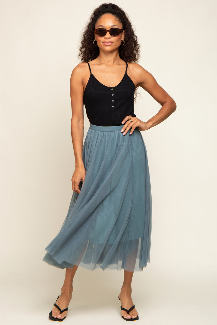woman wearing a black ribbed tank top and a sheer teal maxi skirt, accessorized with sunglasses and sandals