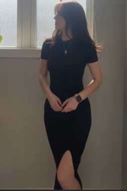 woman wearing a black fitted dress with a side slit, standing by a window with soft lighting