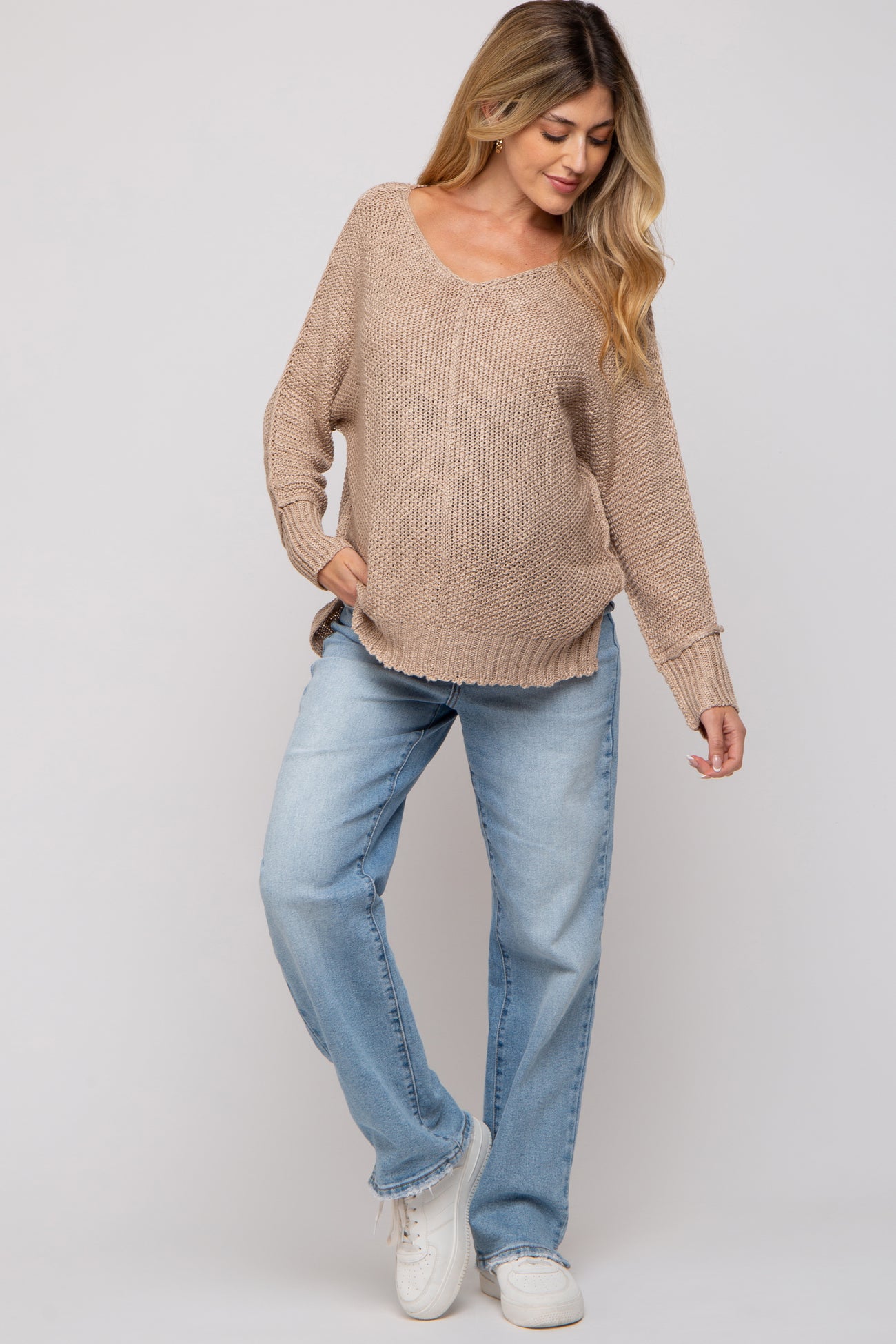 woman wearing a beige oversized knit maternity sweater and light wash jeans, posing with one foot forward