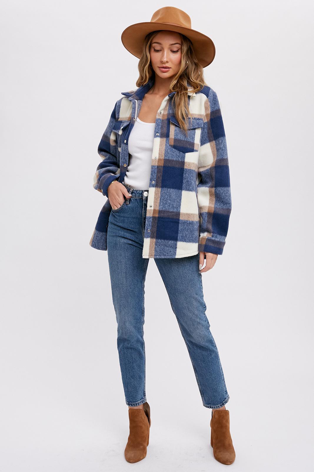 woman wearing a navy blue and cream plaid shacket with a white tank top, blue jeans, and a brown hat