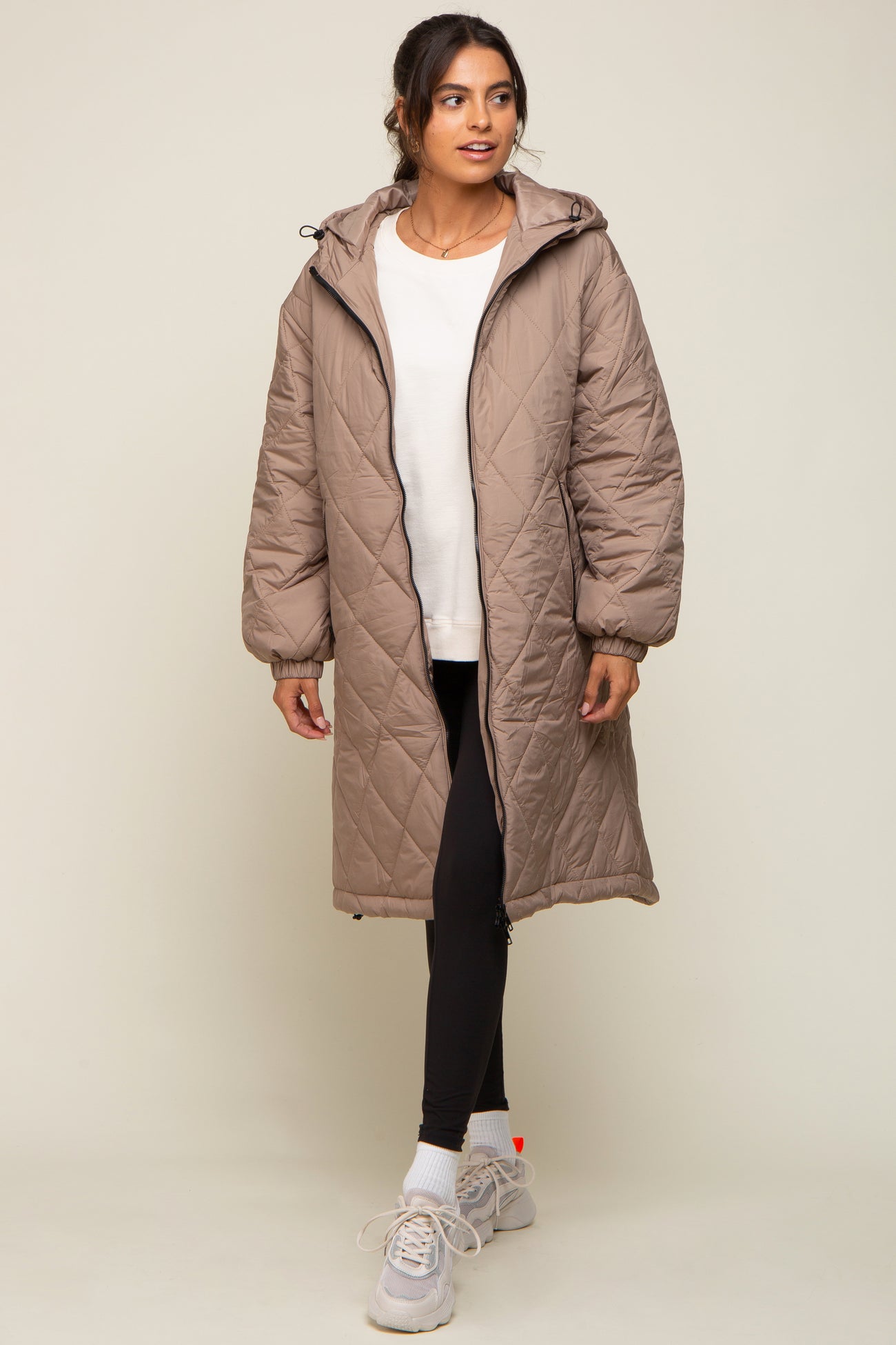 woman wearing a beige padded long coat over a white top and black leggings, paired with gray sneakers
