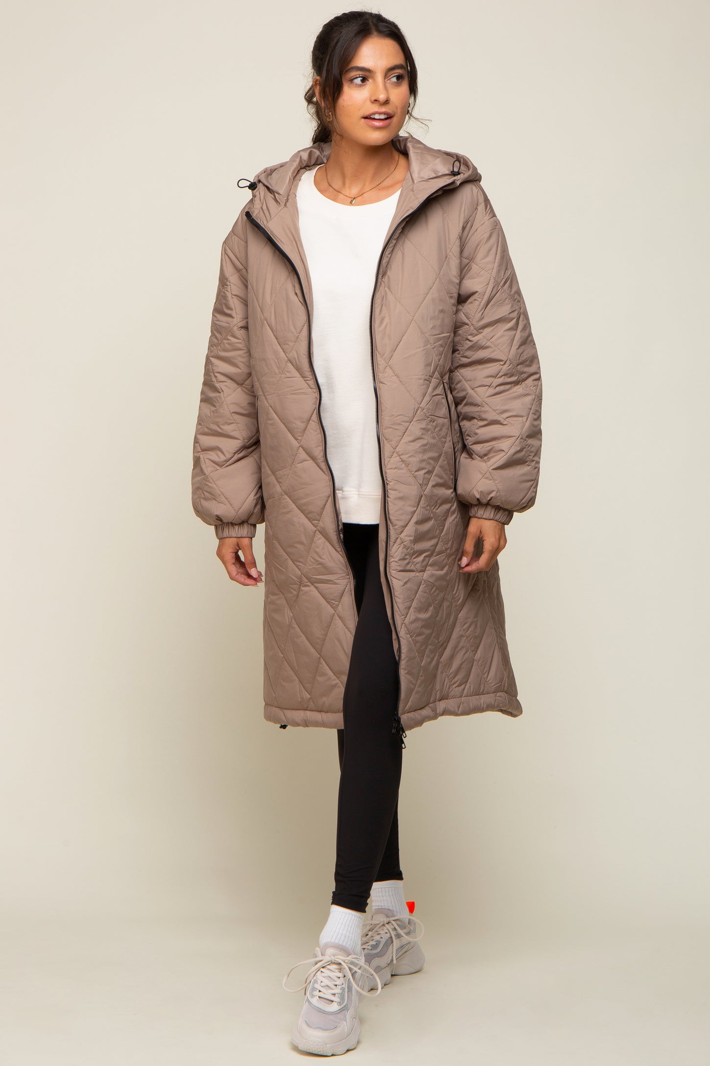 woman wearing a beige padded long coat over a white top and black leggings, paired with gray sneakers