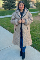 woman wearing a beige long quilted puffer coat, black sweater, and blue jeans, standing outdoors in a park