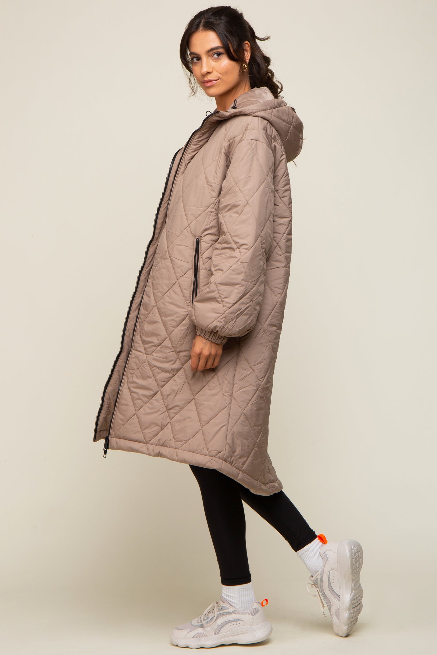 Woman wearing a long taupe quilted puffer coat with a hood, black leggings, and white sneakers