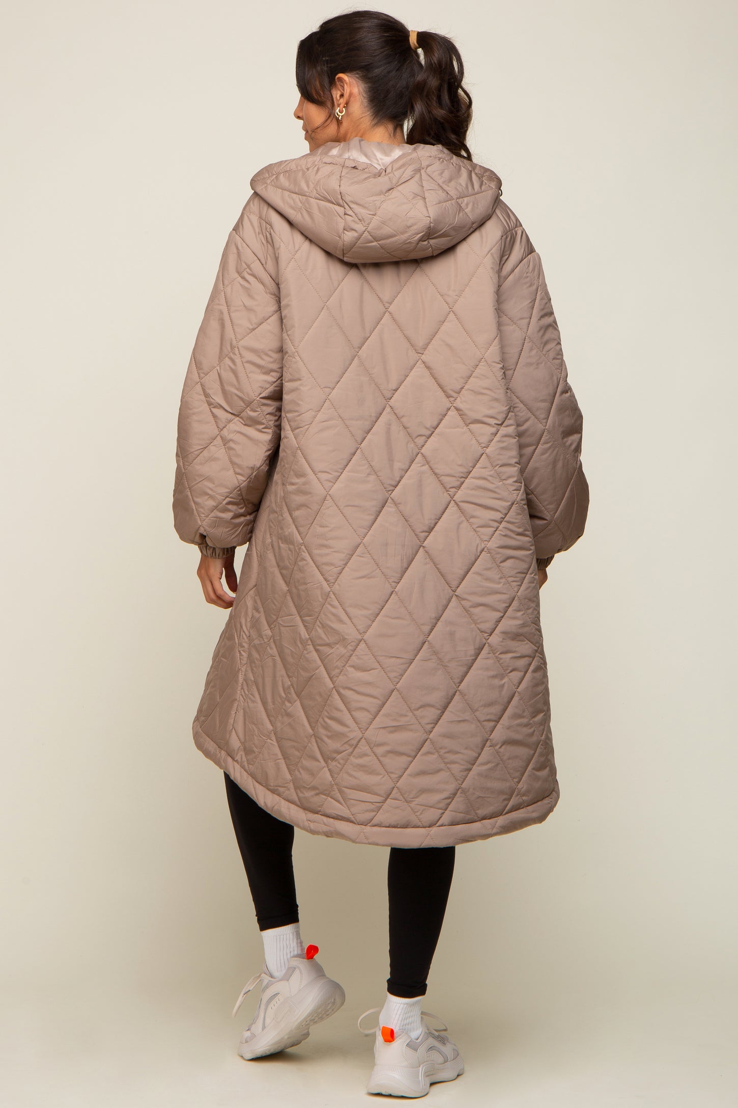 woman wearing a long beige quilted coat with a hood, showing the back view and paired with black leggings and sneakers