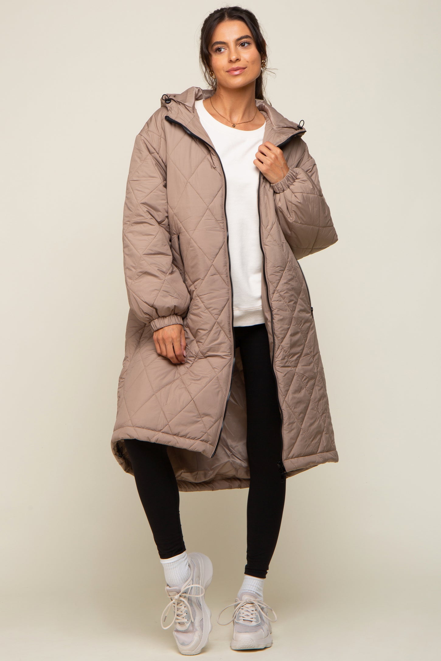 woman wearing a long beige quilted coat over a white sweater and black leggings, paired with white sneakers