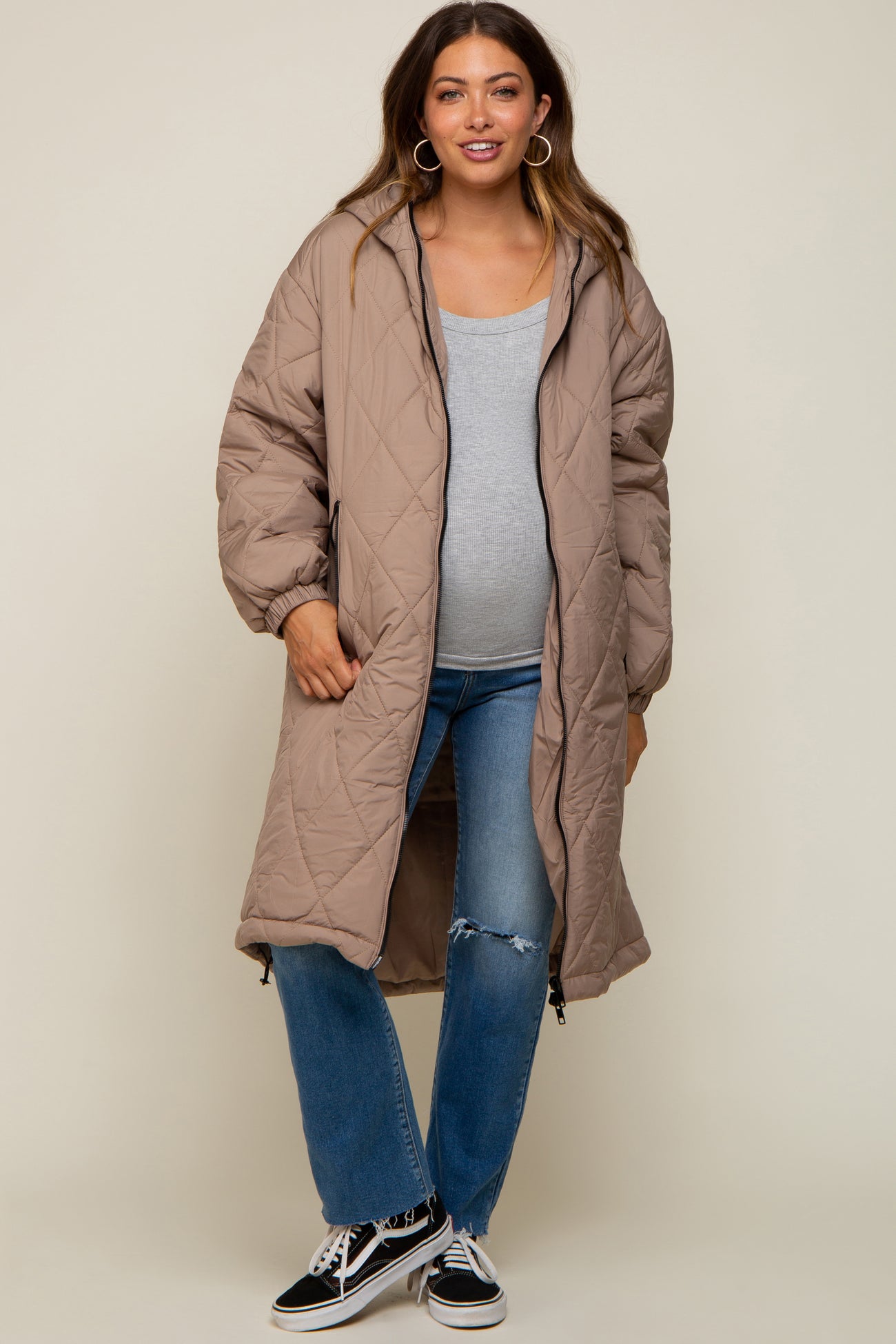 woman wearing a beige quilted maternity coat over a grey top and distressed jeans, showing her baby bump