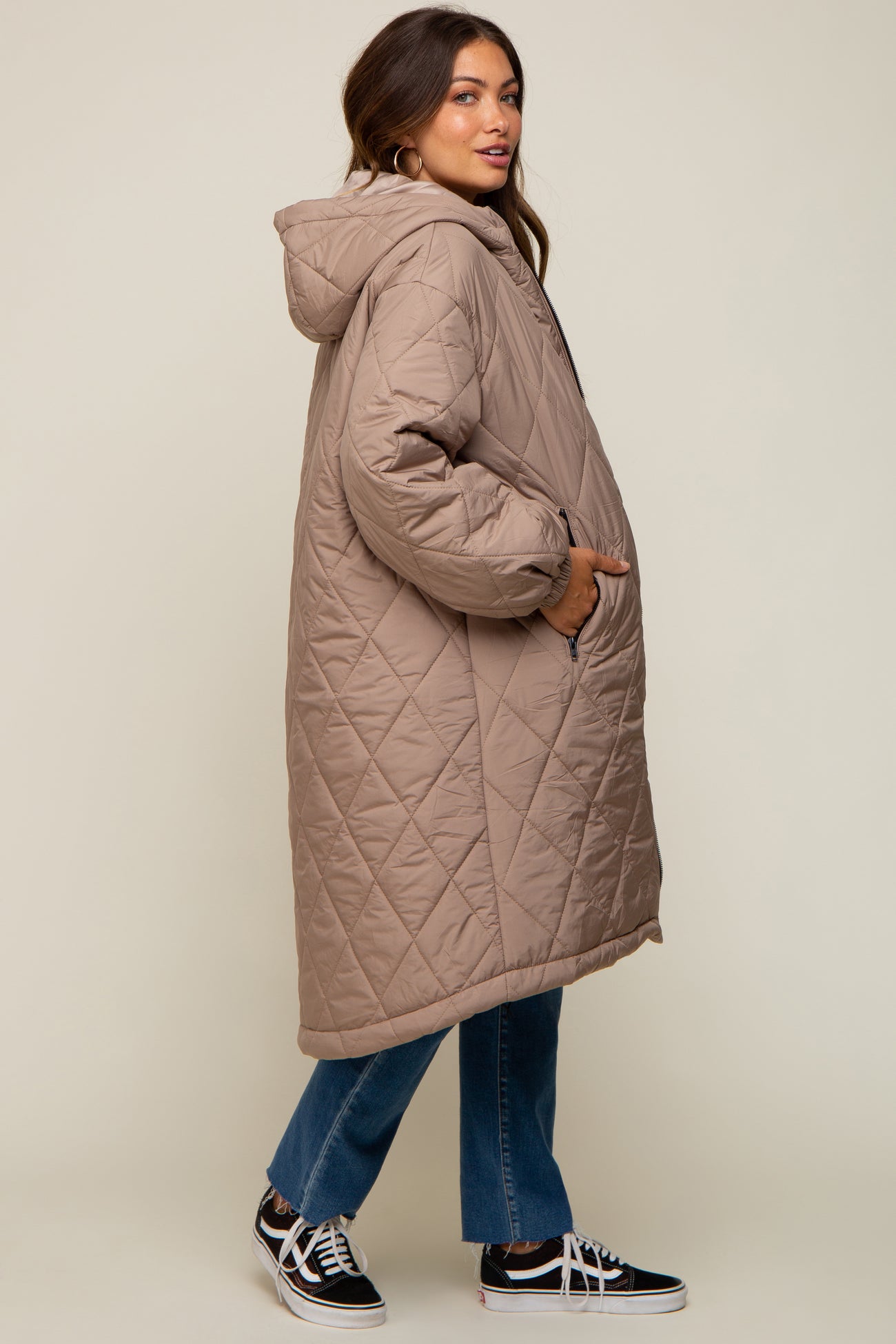 woman wearing a beige quilted maternity coat with a hood and side pockets, styled with jeans and sneakers