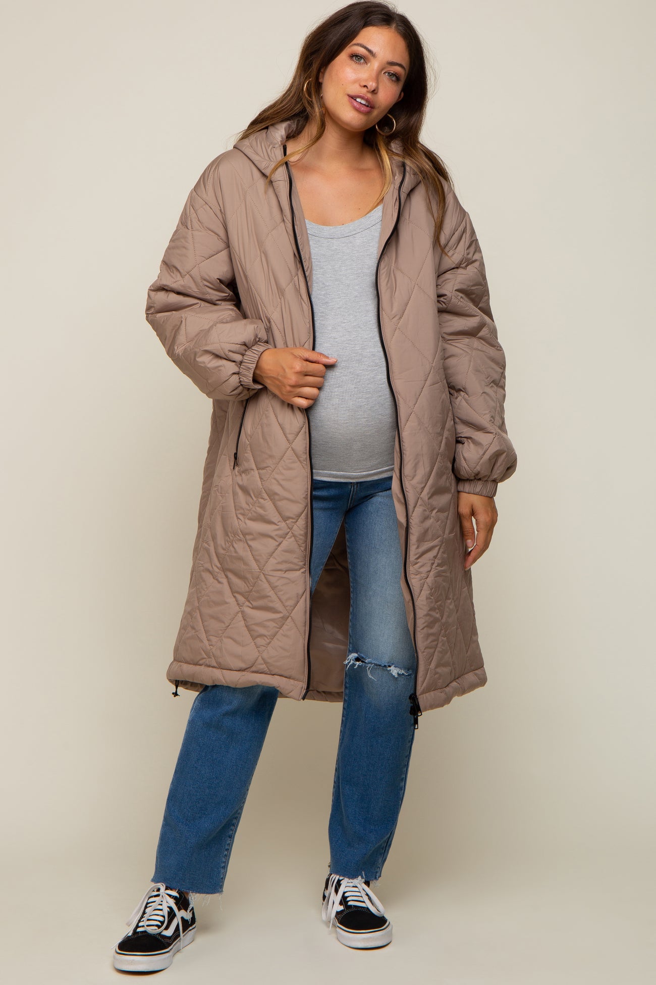 woman wearing a light brown maternity puffer coat over a gray top and blue jeans, showing her baby bump