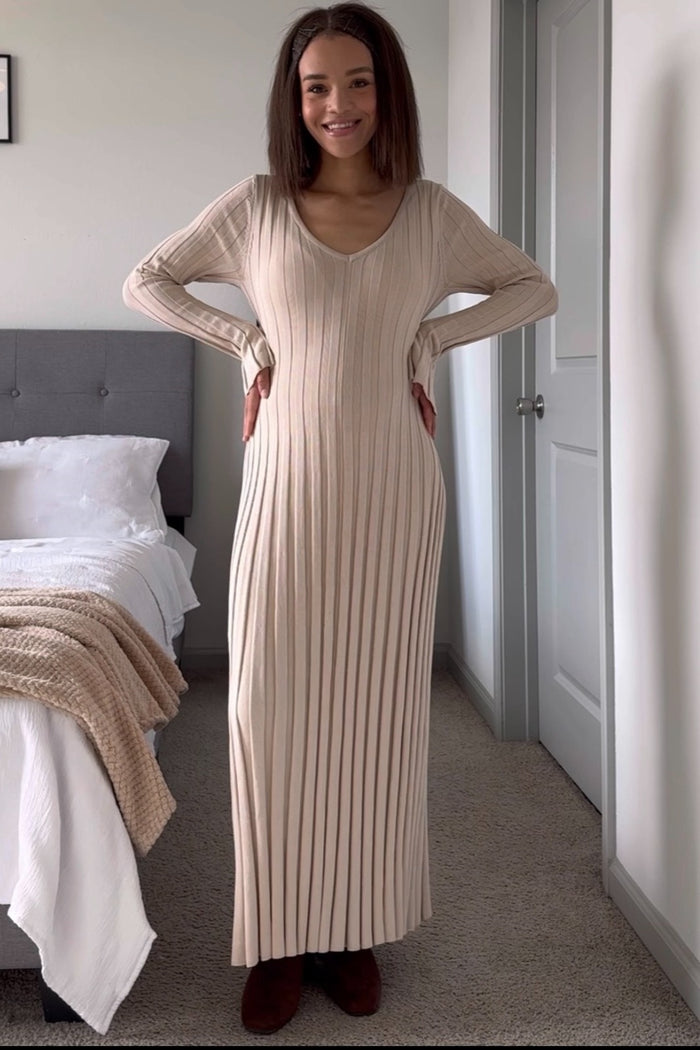 woman wearing a beige ribbed maxi dress with a deep v-neck and long sleeves, standing in a bedroom setting