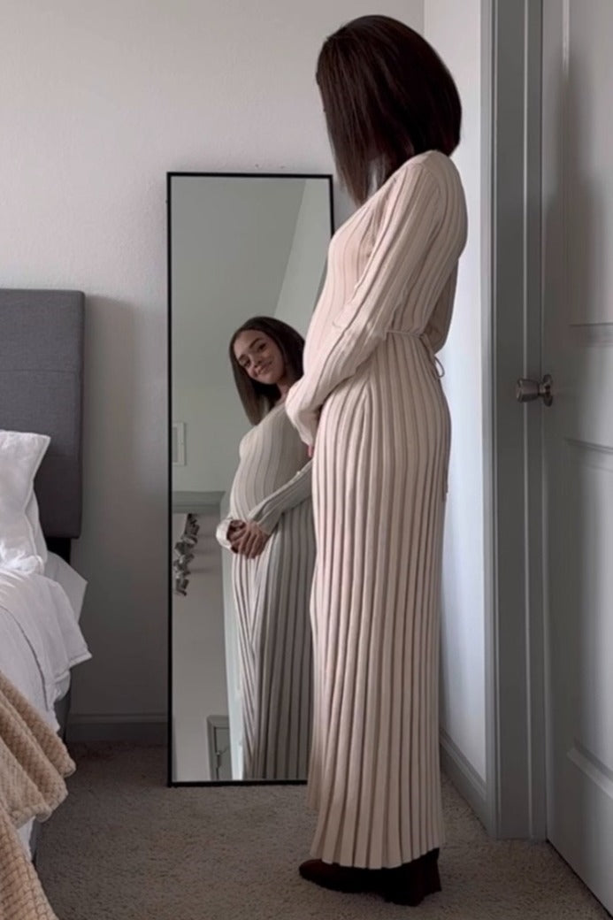 woman wearing a beige ribbed maternity dress, smiling and holding her baby bump while looking in a mirror