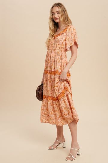 woman wearing a light orange floral maxi dress with short sleeves and tiered detailing, standing with one hand in pocket