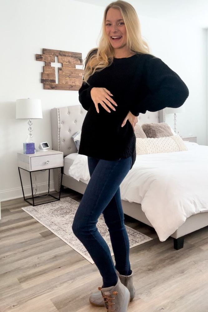 woman wearing a black oversized maternity sweater and dark skinny jeans, holding her baby bump in a cozy bedroom setting