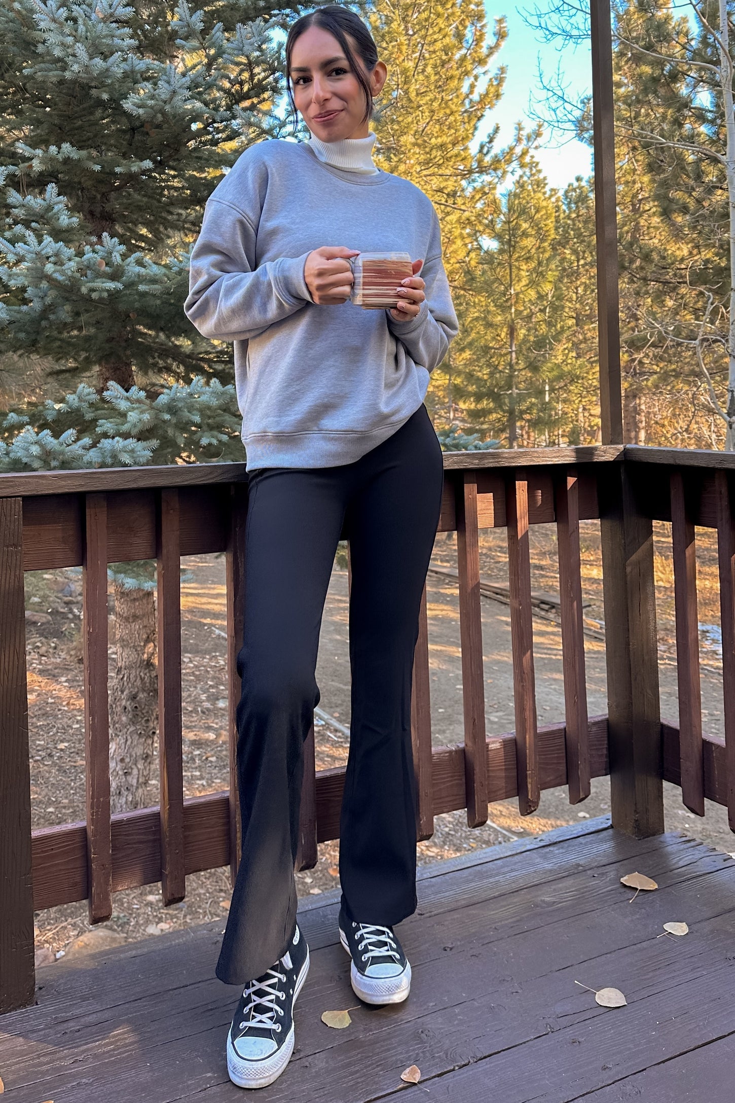 woman wearing a gray sweatshirt and black flared pants, holding a mug while standing on a wooden deck in a forest setting