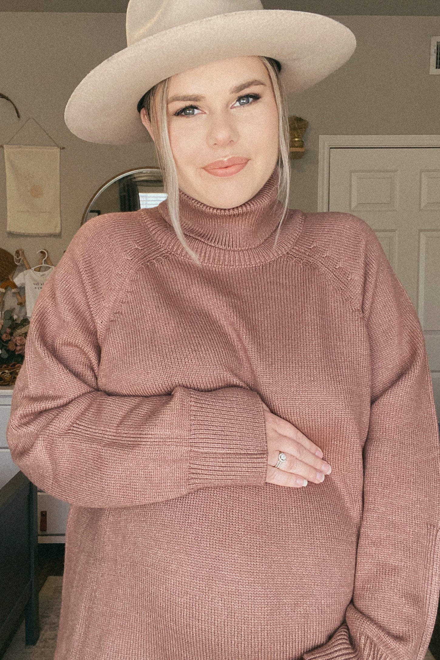 woman wearing a dusty rose maternity turtleneck sweater and holding her baby bump, with a wide-brimmed hat
