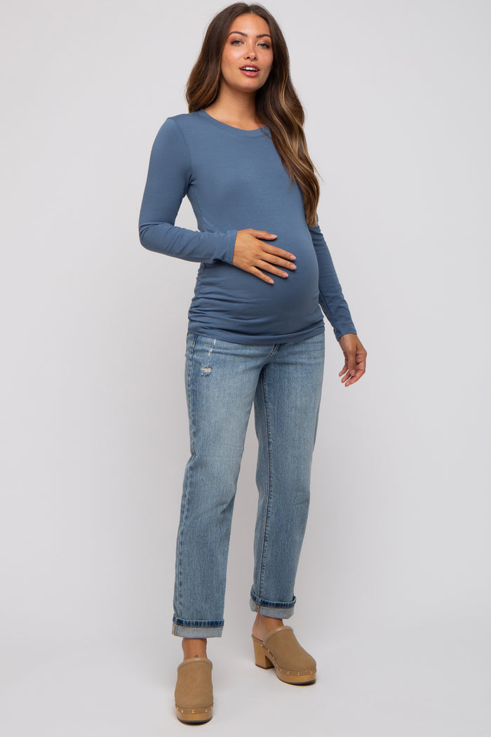 woman wearing a blue long sleeve maternity top and holding her baby bump, paired with distressed jeans and clogs