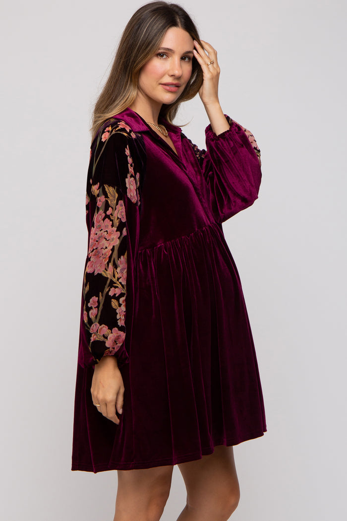 woman wearing a burgundy velvet maternity dress with floral embroidered long sleeves and a flared silhouette, holding her hair back