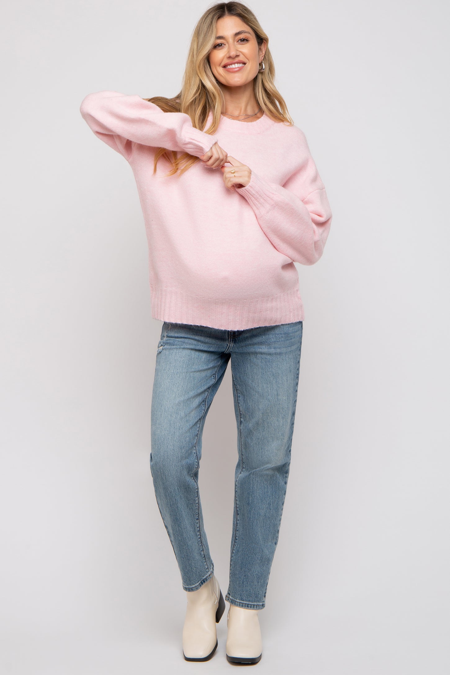 woman wearing a light pink maternity sweater and blue jeans, showing her baby bump while smiling