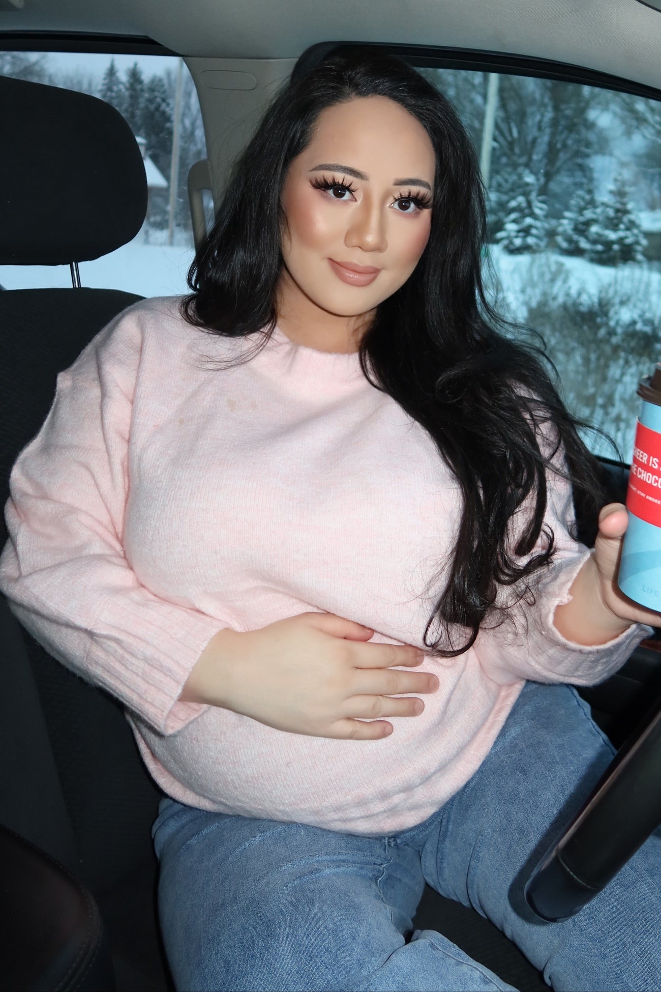 woman wearing a light pink maternity sweater and holding her baby bump while sitting in a car