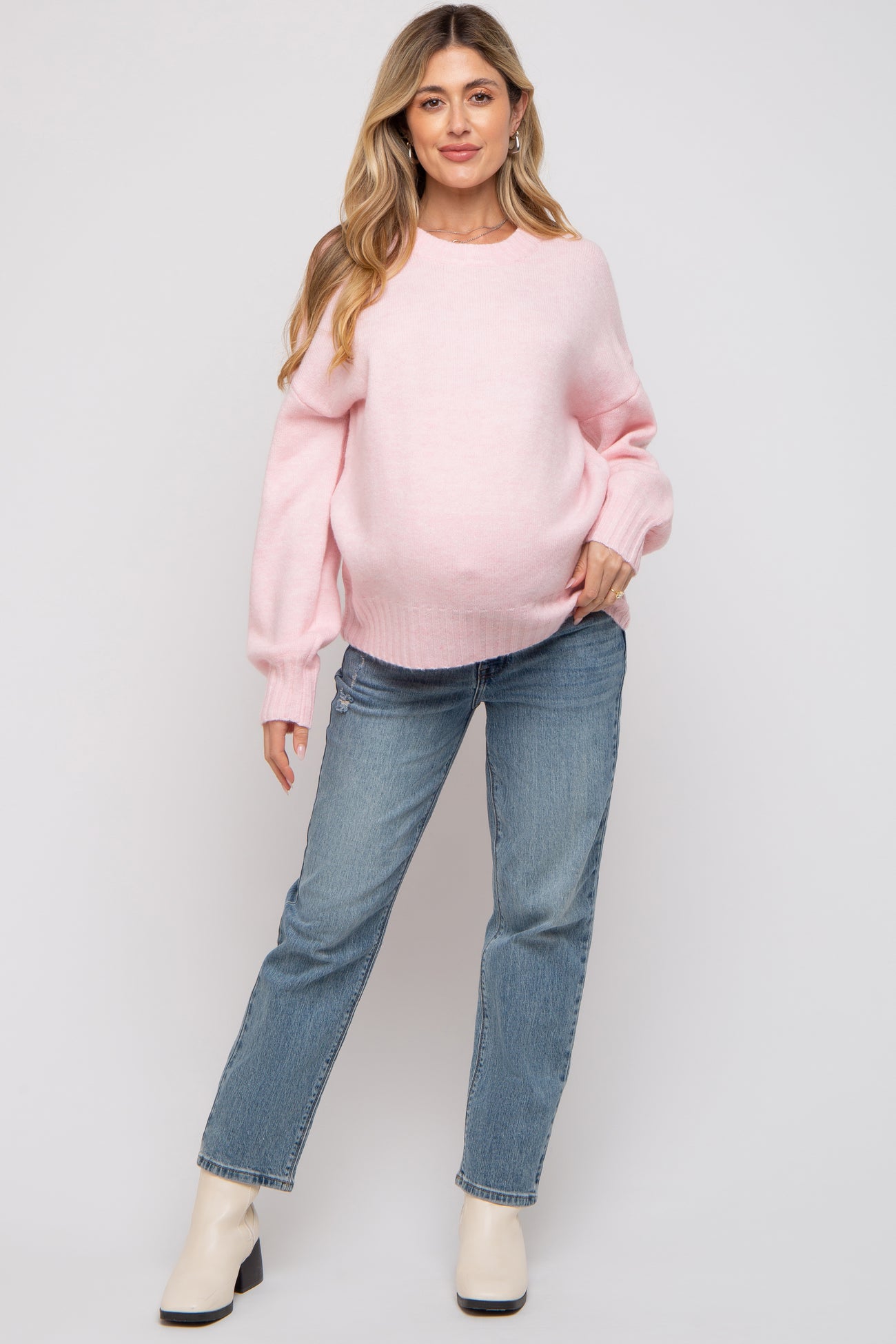 woman wearing a pink maternity sweater and blue jeans, showing her baby bump, standing in a relaxed pose