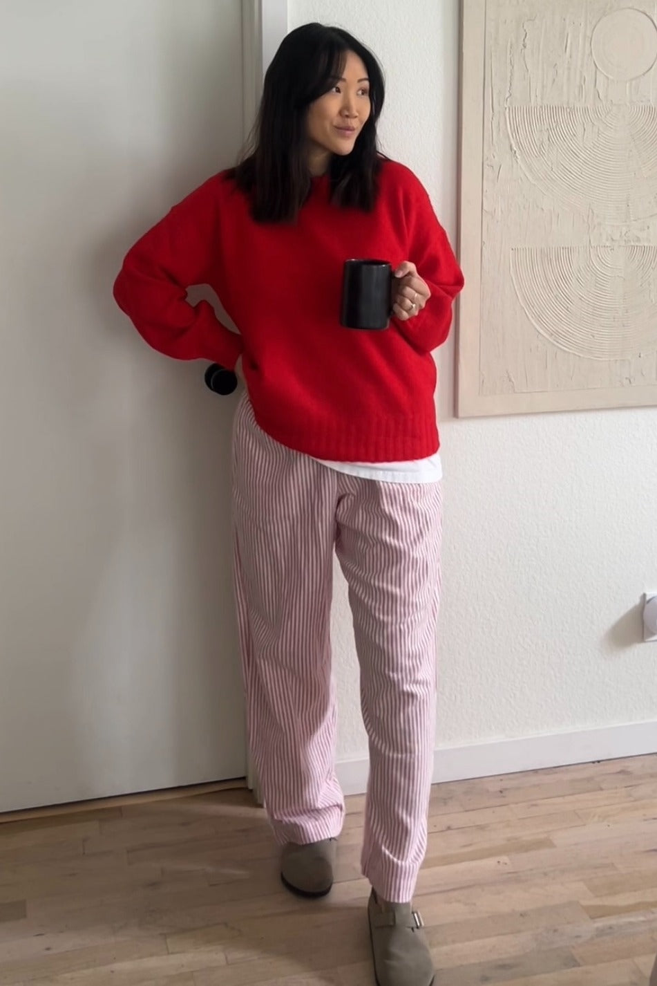 woman wearing a red oversized sweater and pink striped pajama pants, holding a black mug in a cozy setting