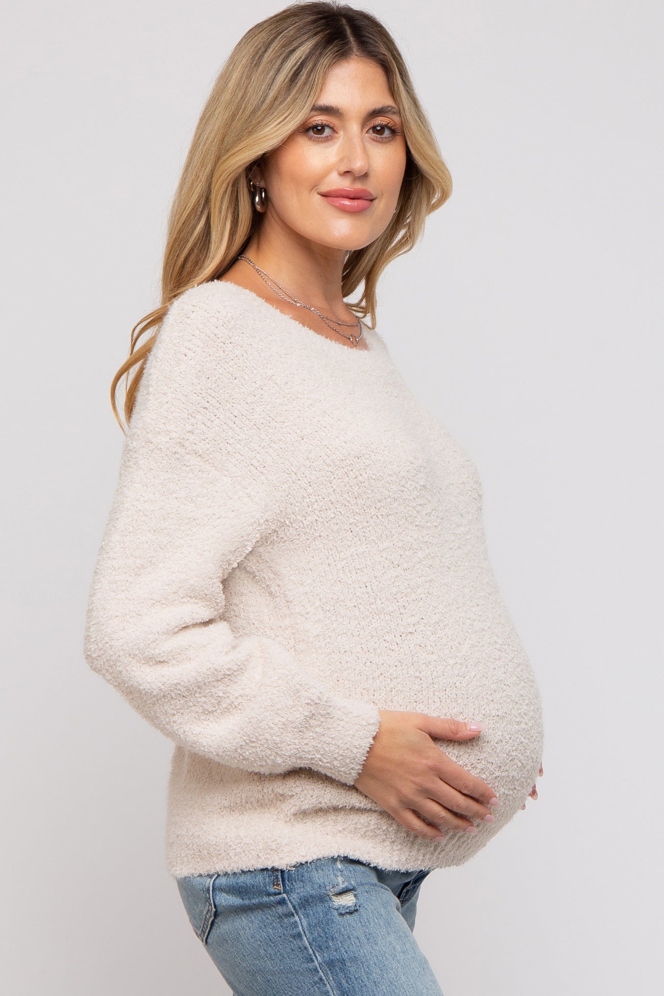 woman wearing a soft beige maternity sweater and cradling her baby bump, paired with light blue jeans