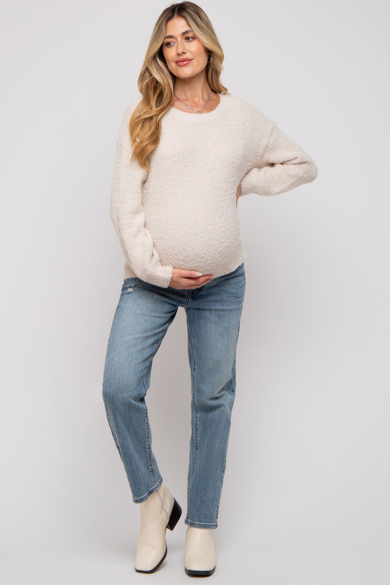 woman wearing a beige maternity sweater and blue distressed jeans, cradling her baby bump