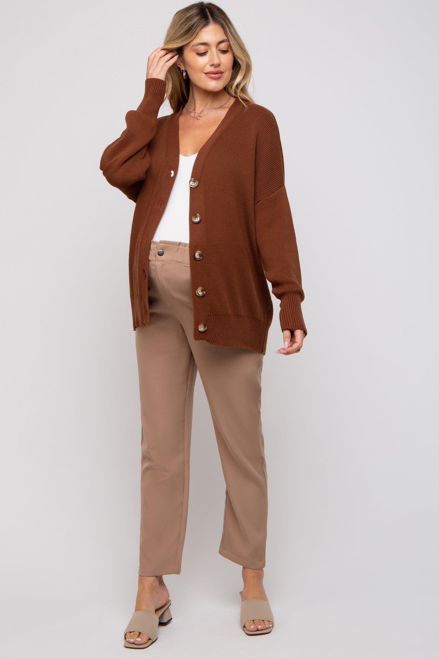 woman wearing a rust brown maternity cardigan over a white top with button details and beige maternity pants