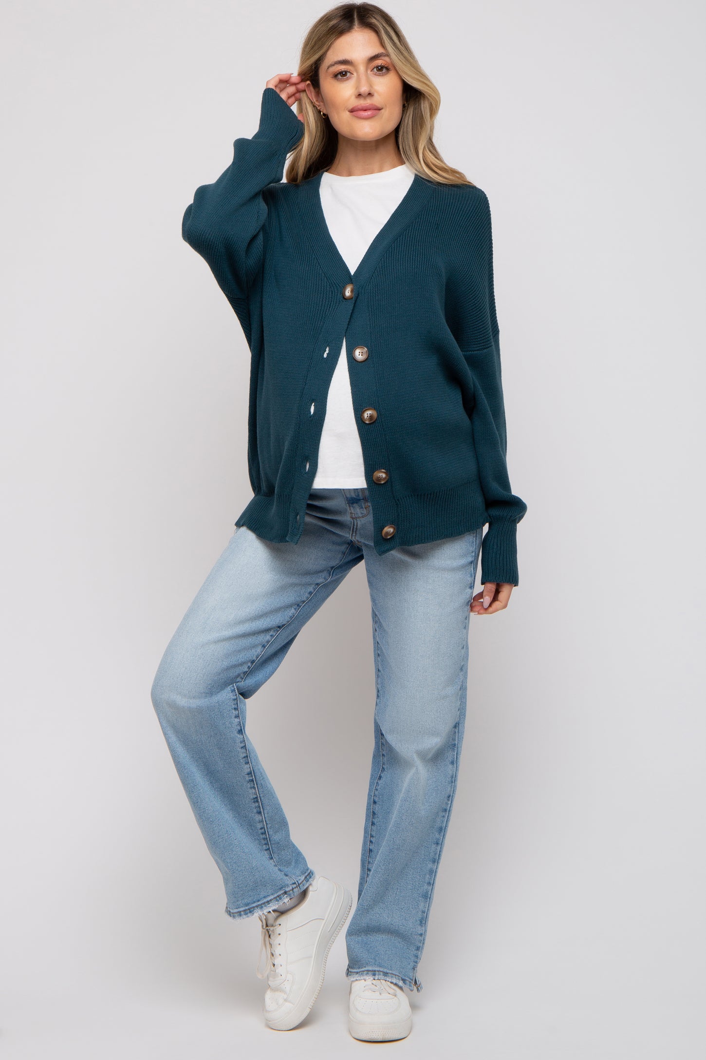 woman wearing a teal maternity cardigan over a white top and light blue jeans, standing in a casual pose