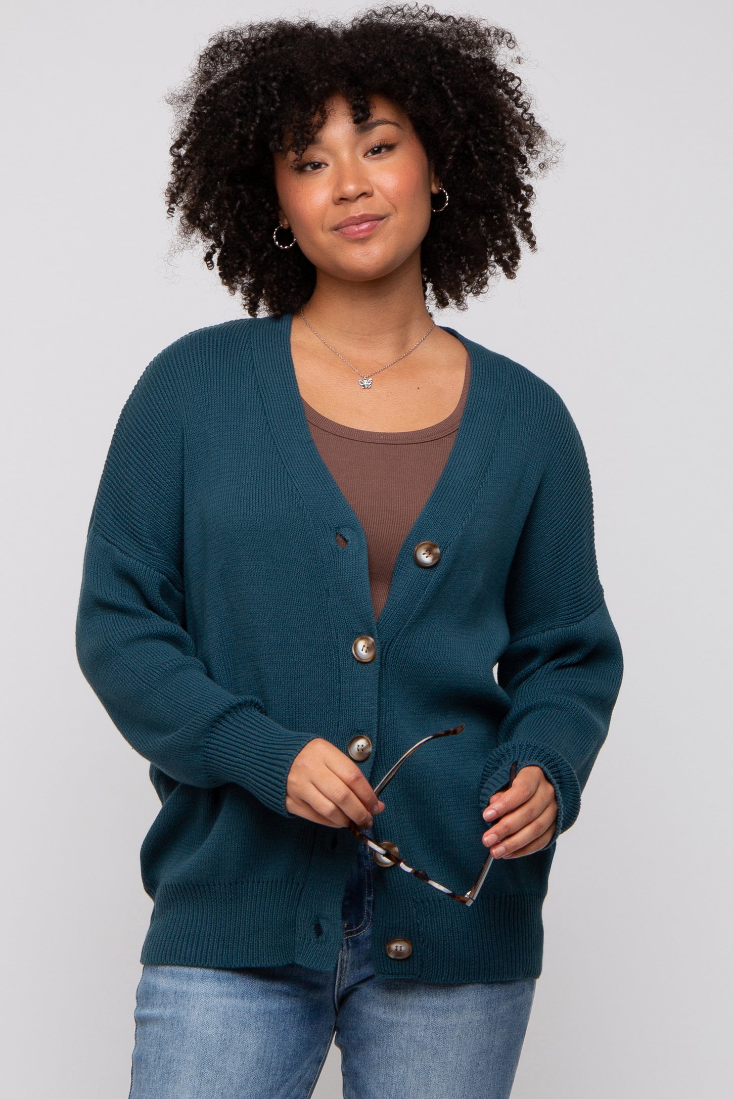 woman wearing a teal button-up cardigan over a brown top and holding sunglasses