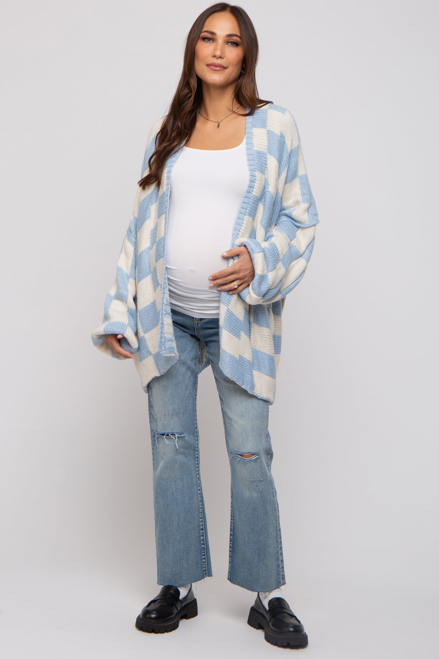woman wearing a light blue and white checkered maternity cardigan and distressed jeans, showing her baby bump