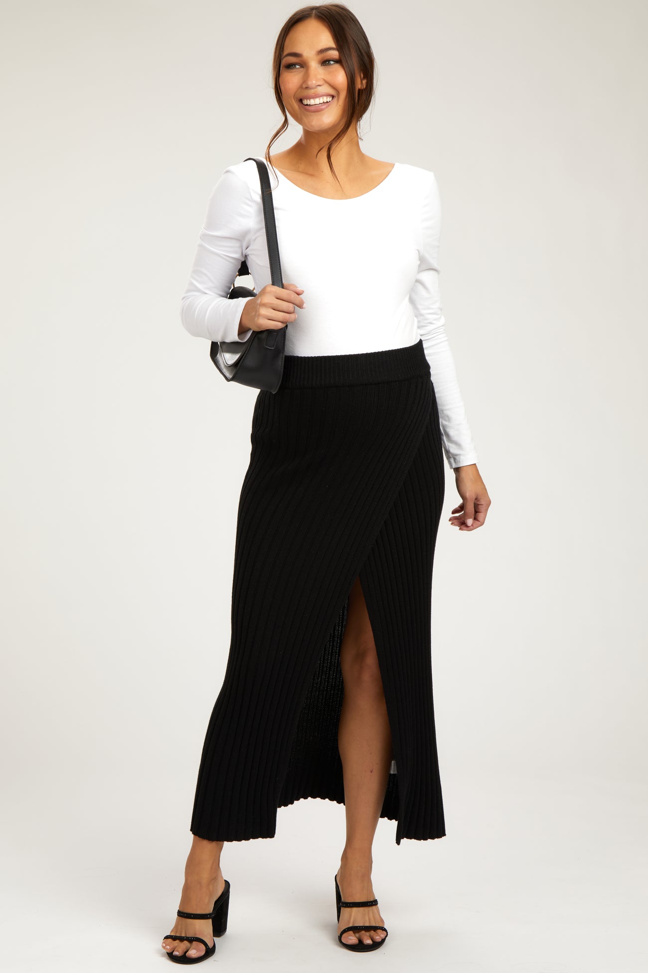 woman wearing a white long sleeve maternity top and a black ribbed maxi skirt with a side slit, holding a bag