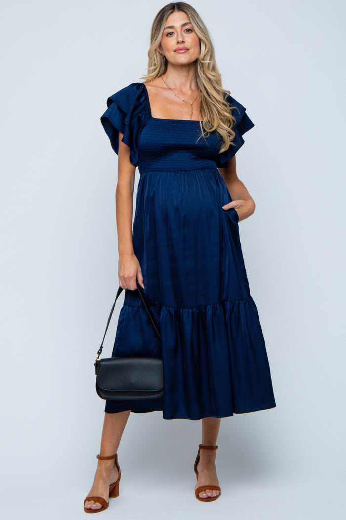 woman wearing a navy blue maternity dress with ruffled sleeves, fitted bodice, and tiered skirt, holding a black handbag