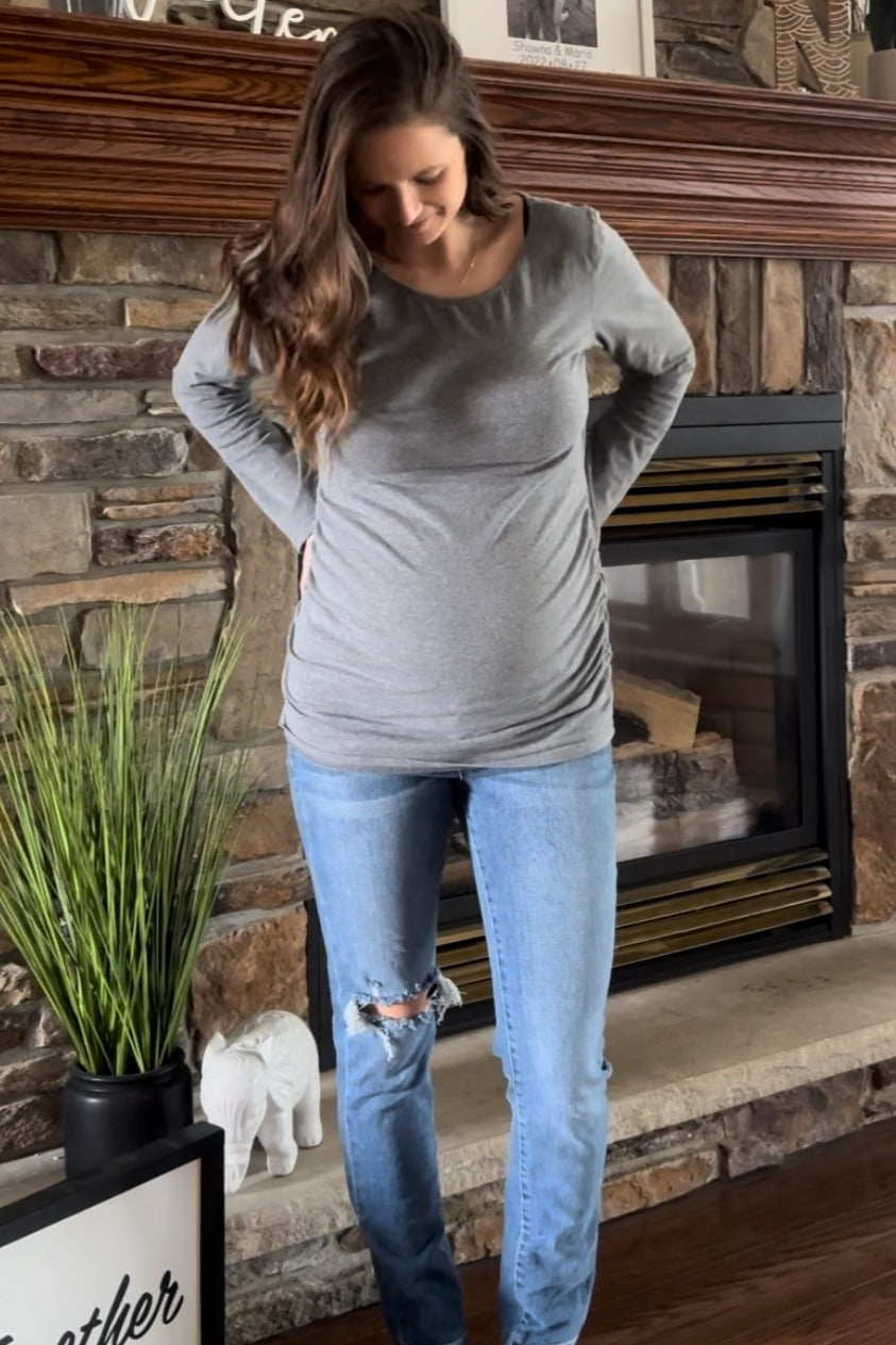 woman wearing a gray long sleeve maternity top with gathered sides, showing her baby bump, paired with ripped blue jeans