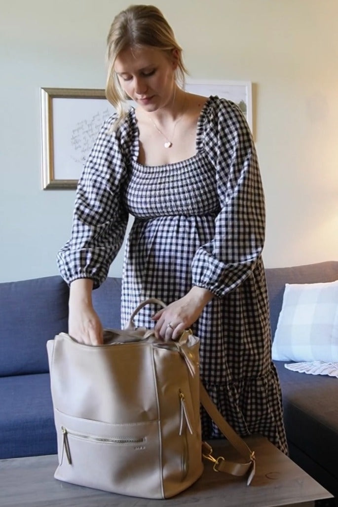 woman wearing a black and white gingham maternity dress and reaching into a beige diaper bag while showing her baby bump