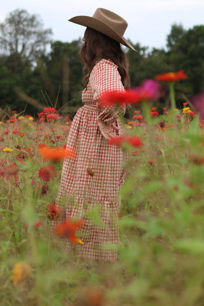 woman wearing a brown gingham maternity dress with a ruffled hem and a wide brim hat, showcasing her baby bump in a flower field