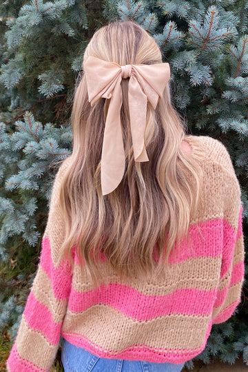 woman wearing a pink and beige striped sweater with a large beige bow in her hair, standing in front of evergreen trees