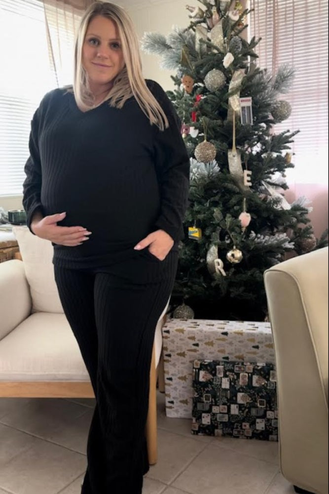 woman wearing a black ribbed maternity loungewear set with a visible baby bump, standing by a decorated Christmas tree