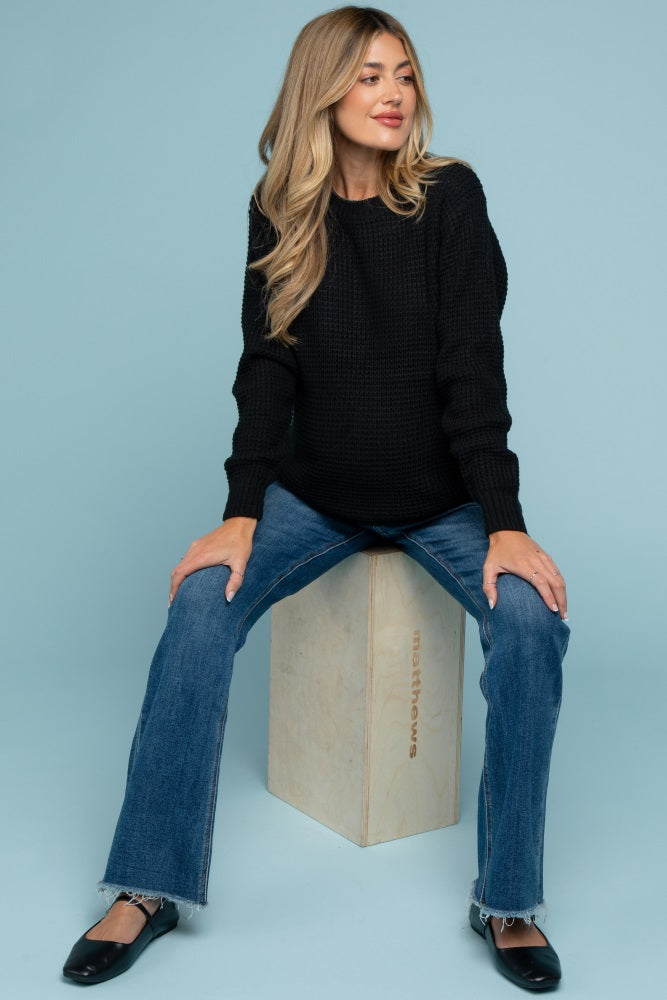 woman wearing a black knitted maternity sweater and blue flared jeans, showing her baby bump while seated on a block